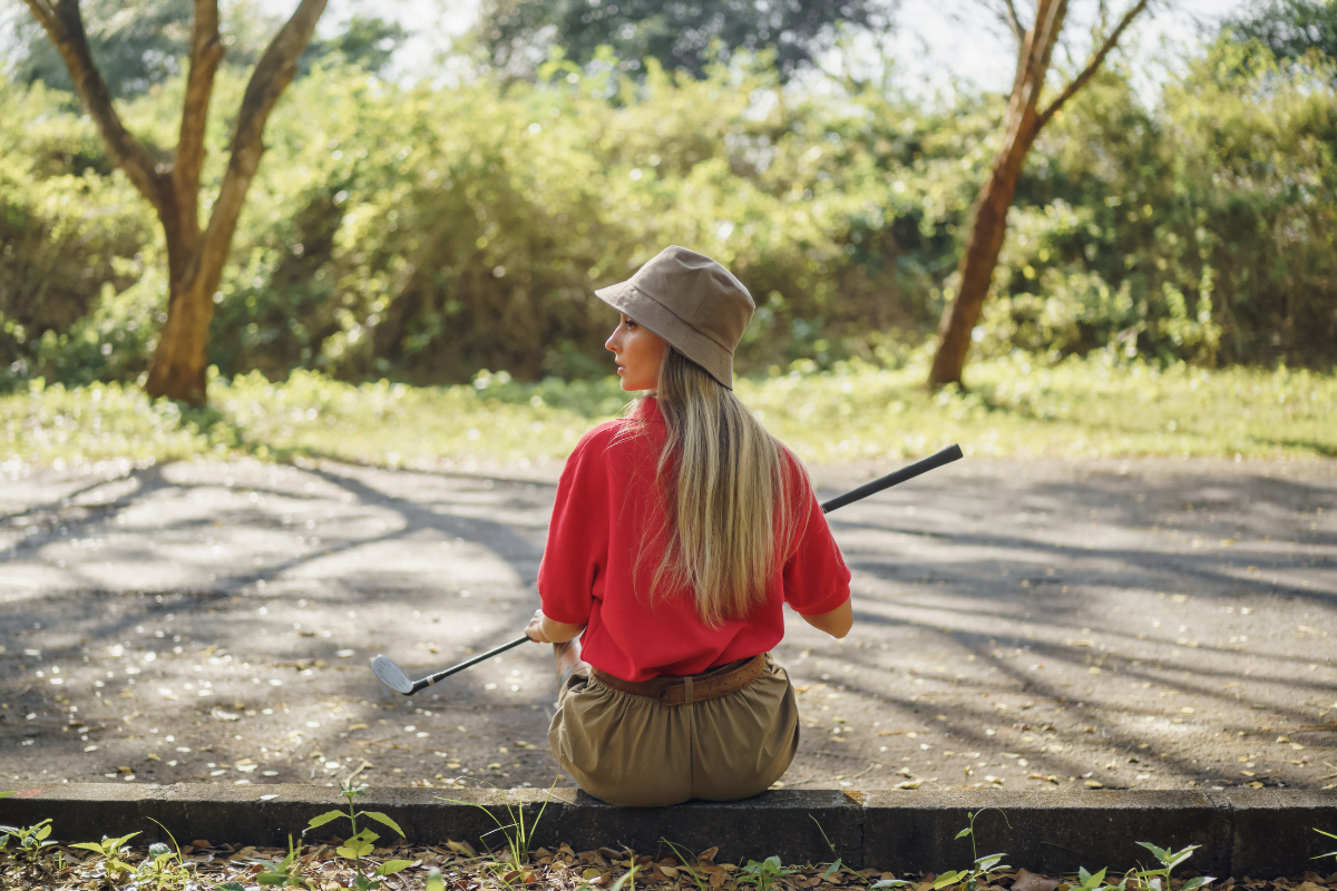 A woman wearing a bucket hat, sitting on the ground with a golf club.