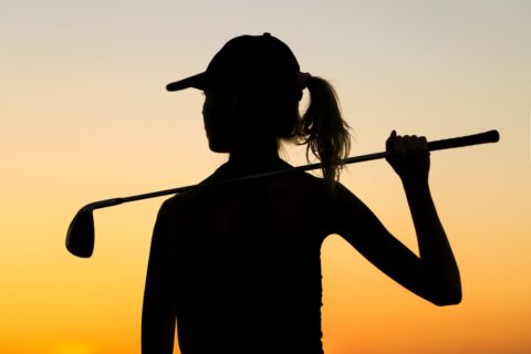 Silhouette of a female golfer, showcasing women's golf fashion, holding a club at sunset.
