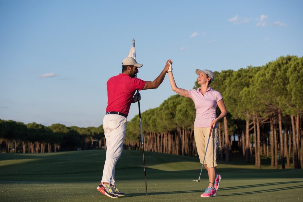 Two golfers high-fiving on a golf course with a flagstick in the background, showcasing women's golf fashion.