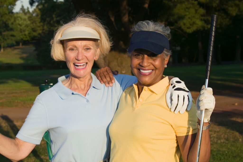 Two senior women smiling on a golf course, one holding a golf club, both showcasing the latest in women's golf fashion.