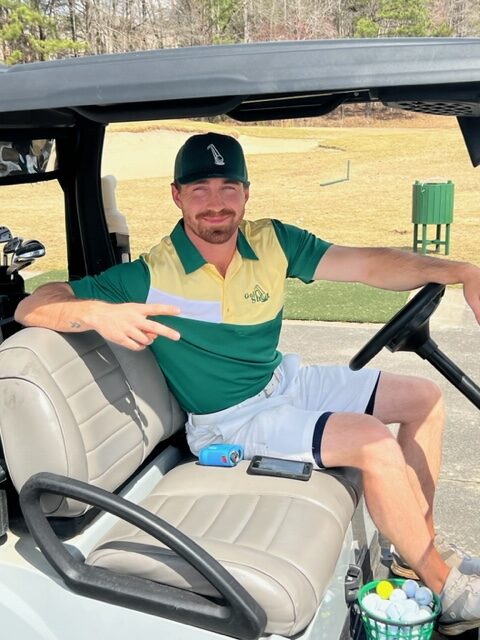 A man sits in a golf cart, holding up a peace sign. He is wearing a green and yellow polo shirt with white shorts and a dark cap. A golf course is visible in the background.