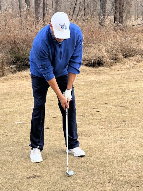 A person in a blue sweater, navy pants, and white cap positions a golf club to hit a ball on a dry grass course.