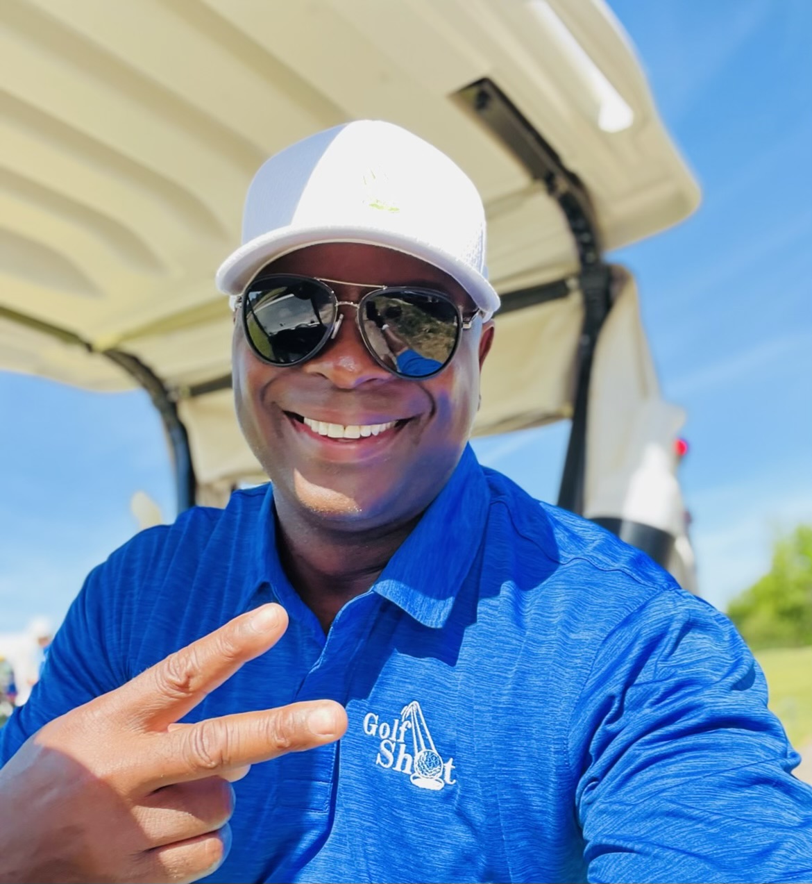 A man wearing sunglasses, a cap, and a blue polo shirt with "Golf Shot" printed on it smiles and gestures a peace sign in front of a golf cart.