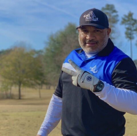 A man in a blue and black polo shirt and a black cap is outdoors, making a peace sign with his right hand while wearing a golf glove, as if proudly celebrating his recent golf shot.