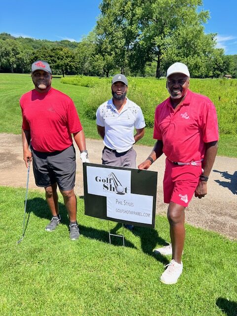 Three men wearing golf attire stand on a golf course next to a sign that reads "About Golf Shot Apparel" in bright daylight. Two of the men are holding golf clubs, with lush greenery in the background.