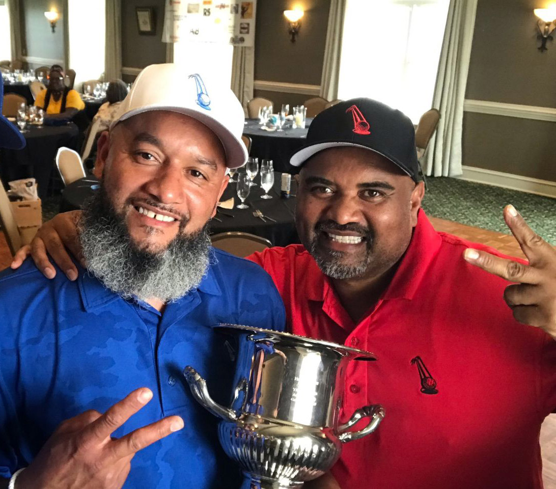 Two men smiling and posing with a silver trophy in a dining hall, both wearing caps and shirts with the same sailboat logo. The man on the left wears blue, the man on the right wears red. .